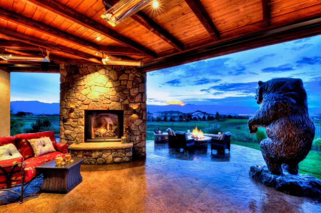 Covered patio in Colorado Springs with a stone fireplace and red seating overlooks an open area at sunset. A large bear sculpture stands near a crackling fire pit.