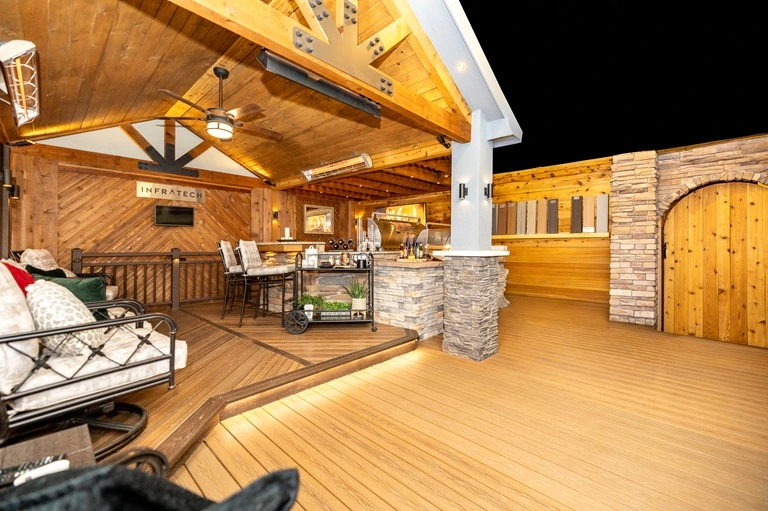A showroom in Englewood that shows a spacious wooden patio featuring cozy seating, a dining area with high chairs, and a stone barbecue area. Warm lighting creates an inviting atmosphere.