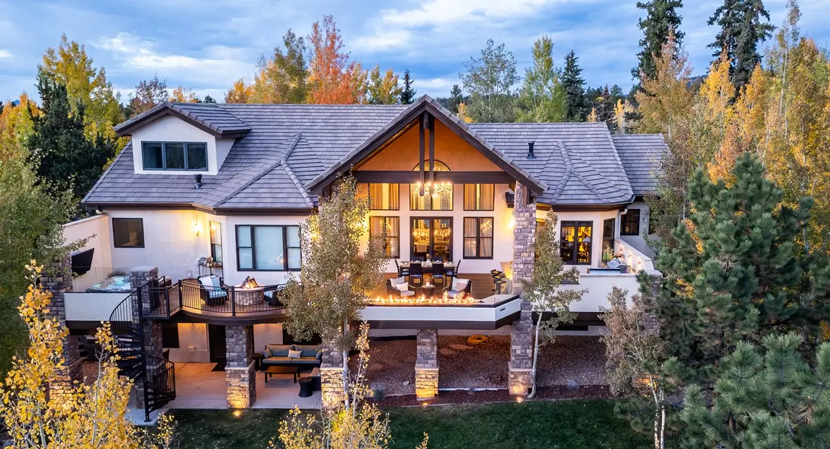 A charming, two-story house in Colorado surrounded by autumn trees, warmly lit at dusk. Large windows, balcony, and soft exterior lights create a cozy, inviting atmosphere.