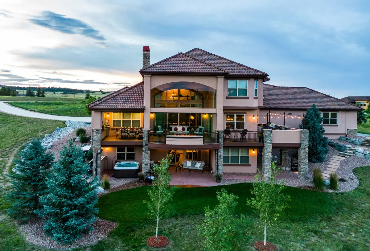 Elegant two-story house with large windows and a custom deck by Mosaic Outdoor Living, surrounded by lush green lawn and trees, set against a serene evening sky.
