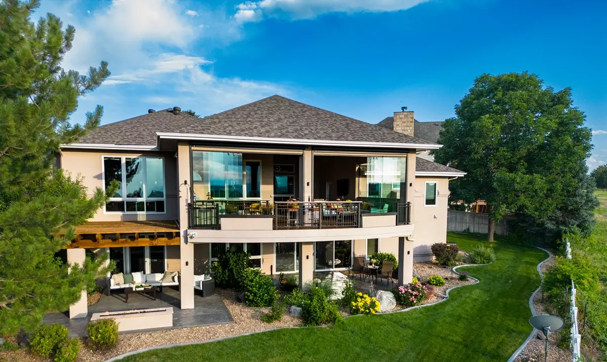 A modern two-story house with large glass windows, a spacious custom deck by Mosaic Outdoor Living, and lush greenery. The scene conveys tranquility under a bright blue sky.