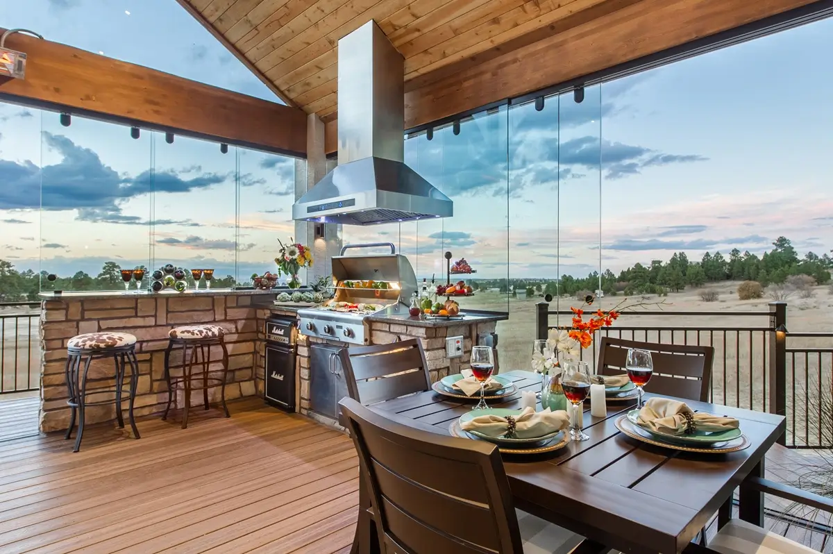 A custom deck by Mosaic Outdoor Living with a modern outdoor kitchen and dining area. A grill is on the left, and a table set for four with wine glasses is on the right, overlooking a scenic landscape.