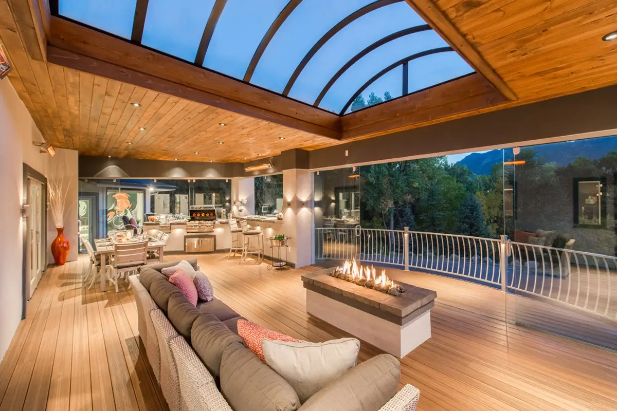 Luxurious custom deck by Mosaic Outdoor Living with a curved glass roof, modern fire pit, plush couch, and dining area. Warm lighting enhances the elegant, cozy ambiance.