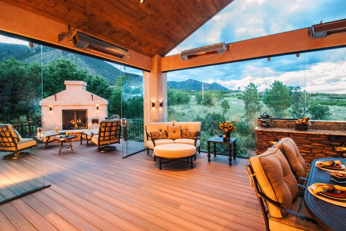 Luxurious custom deck by Mosaic Outdoor Living with cozy seating, fireplace, and dining area. Glass walls reveal a scenic mountain view, creating a warm, inviting atmosphere.